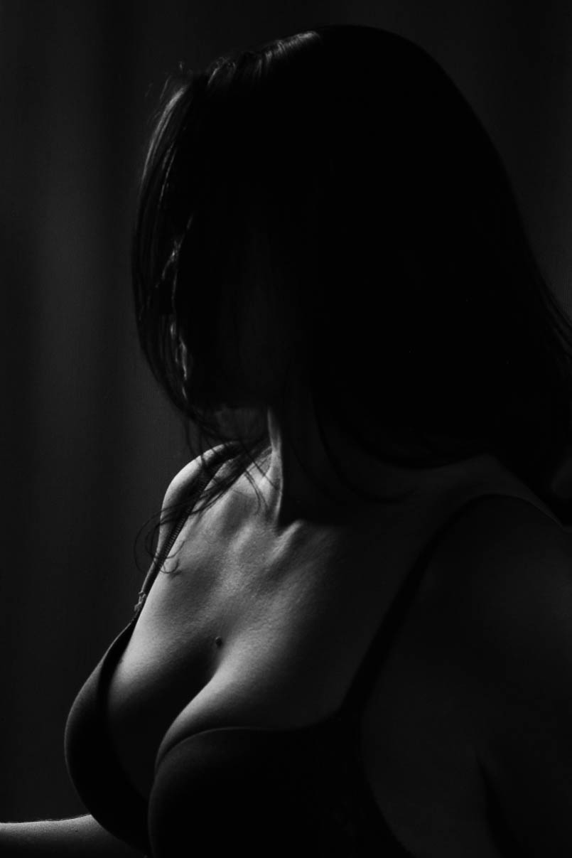 Low-key black and white photograph of a woman looking back out of frame, her face partially obscured by dark hair. A soft shaft of light highlights her chest, neck, shoulder, and bra, creating a moody and intimate atmosphere.